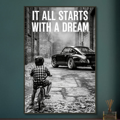 Kleiner Junge auf dem Fahrrad blickt auf einen Porsche 911 mit dem Motivationsspruch "It All Starts With A Dream" auf einem Poster im Metallrahmen