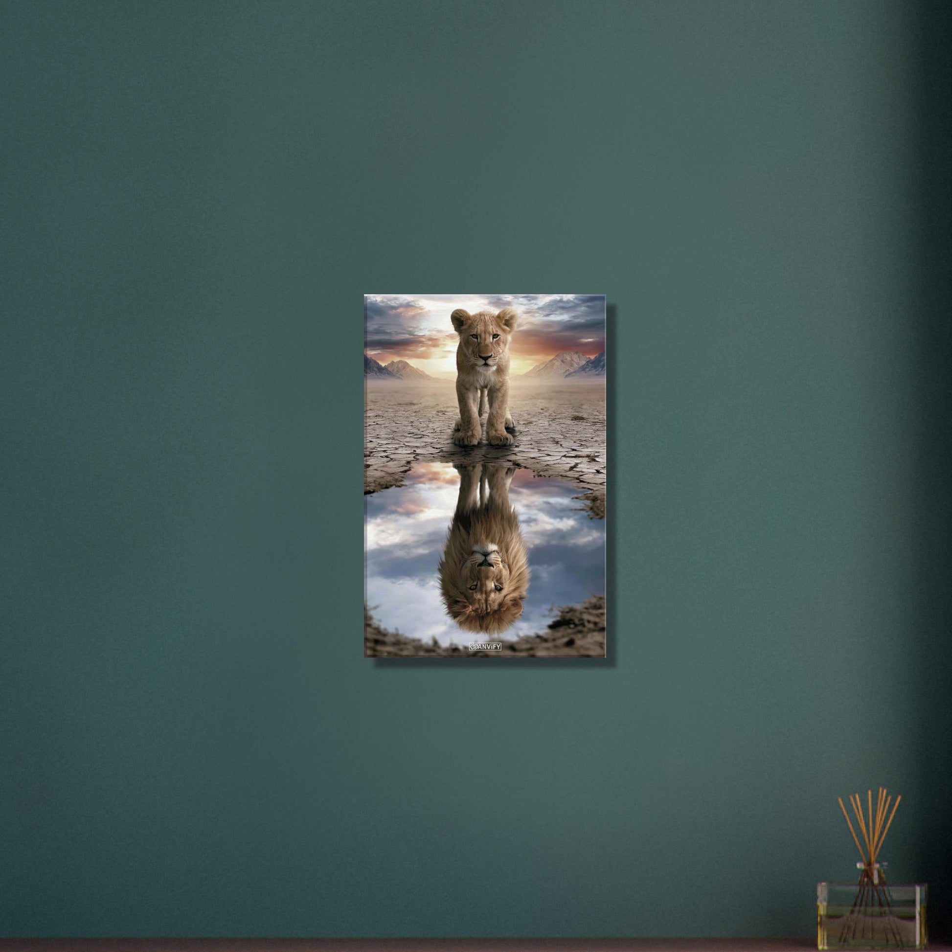 Kleiner Löwe steht vor einer Wasserpfütze, in der sich ein großer Löwe mit Mähne spiegelt – vor Bergpanorama und Sonnenuntergang auf einer Leinwand