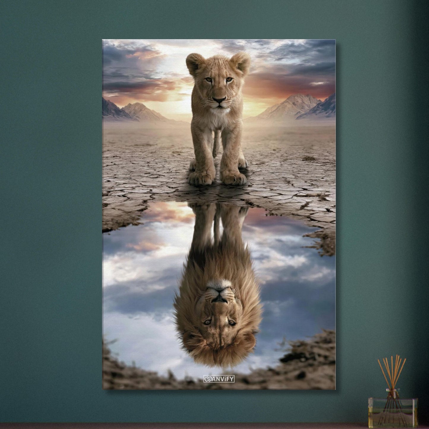 Kleiner Löwe steht vor einer Wasserpfütze, in der sich ein großer Löwe mit Mähne spiegelt – vor Bergpanorama und Sonnenuntergang auf einer Leinwand