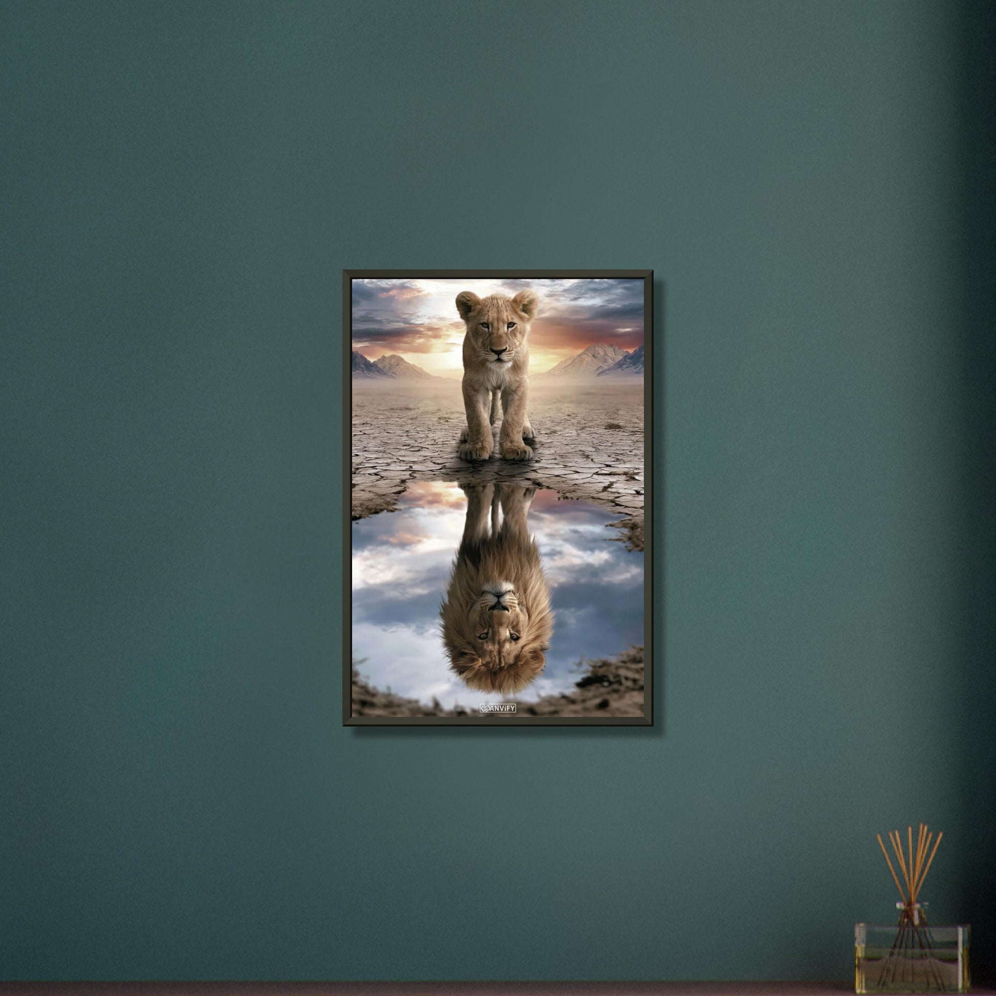 Kleiner Löwe steht vor einer Wasserpfütze, in der sich ein großer Löwe mit Mähne spiegelt – vor Bergpanorama und Sonnenuntergang auf einem Poster im Metallrahmen