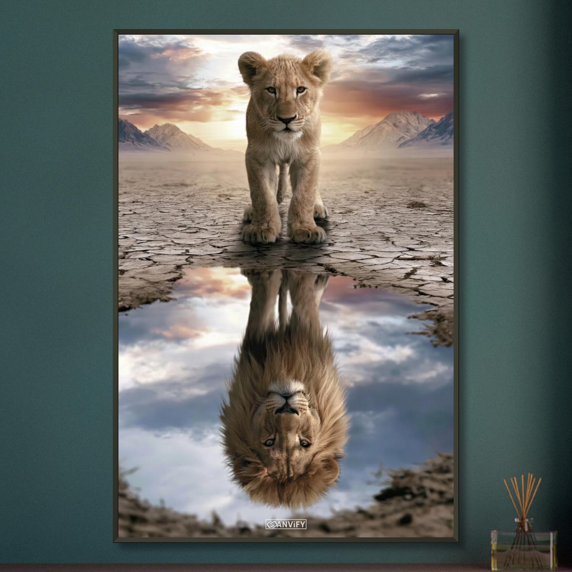 Kleiner Löwe steht vor einer Wasserpfütze, in der sich ein großer Löwe mit Mähne spiegelt – vor Bergpanorama und Sonnenuntergang auf einem Poster im Metallrahmen